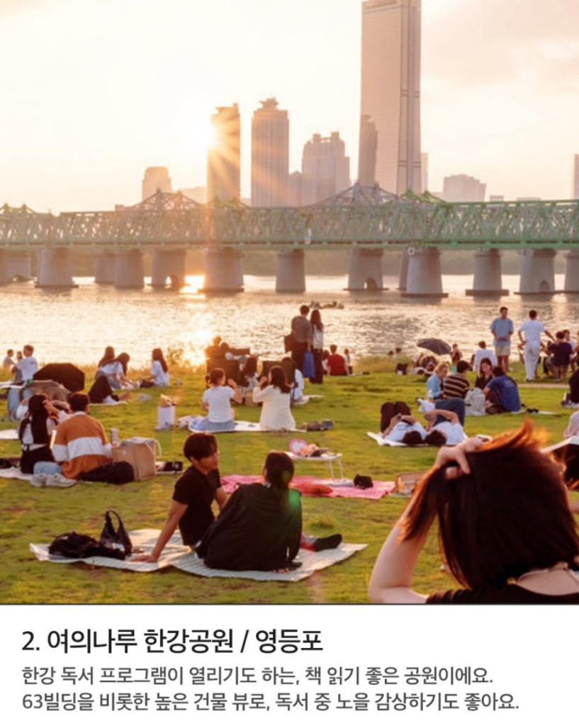 책읽기좋은공원 여의나루 한강공원 석양 무렵 한강공원 잔디밭에 많은 사람들이 돗자리를 펴고 앉아 있다. 다리와 고층 빌딩이 배경에 보인다.